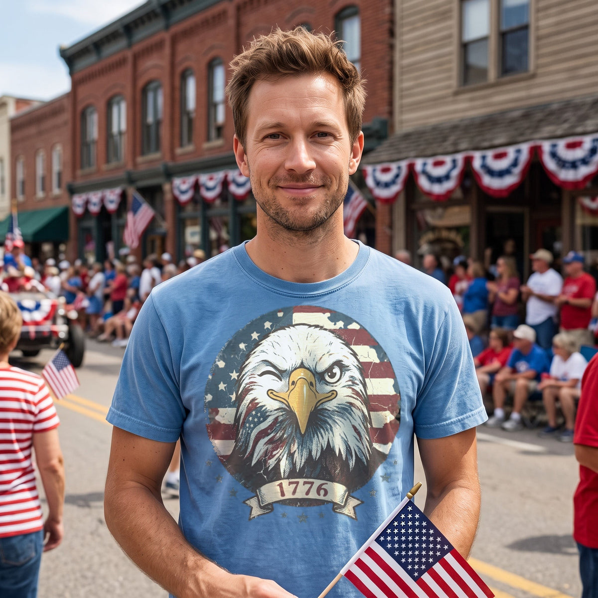 Man in a light blue tee holding a small American flag at a small-town Fourth of July street parade