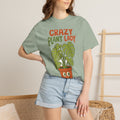 Woman wearing a bay t-shirt printed with the Crazy Plant Lady design, standing beside a wooden ladder and a woven basket against a plain wall