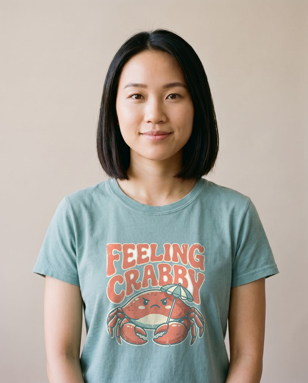 Woman wearing the Feeling Crabby funny crab tee in Bay sage green, retro bubble lettering with grumpy crab under beach umbrella
