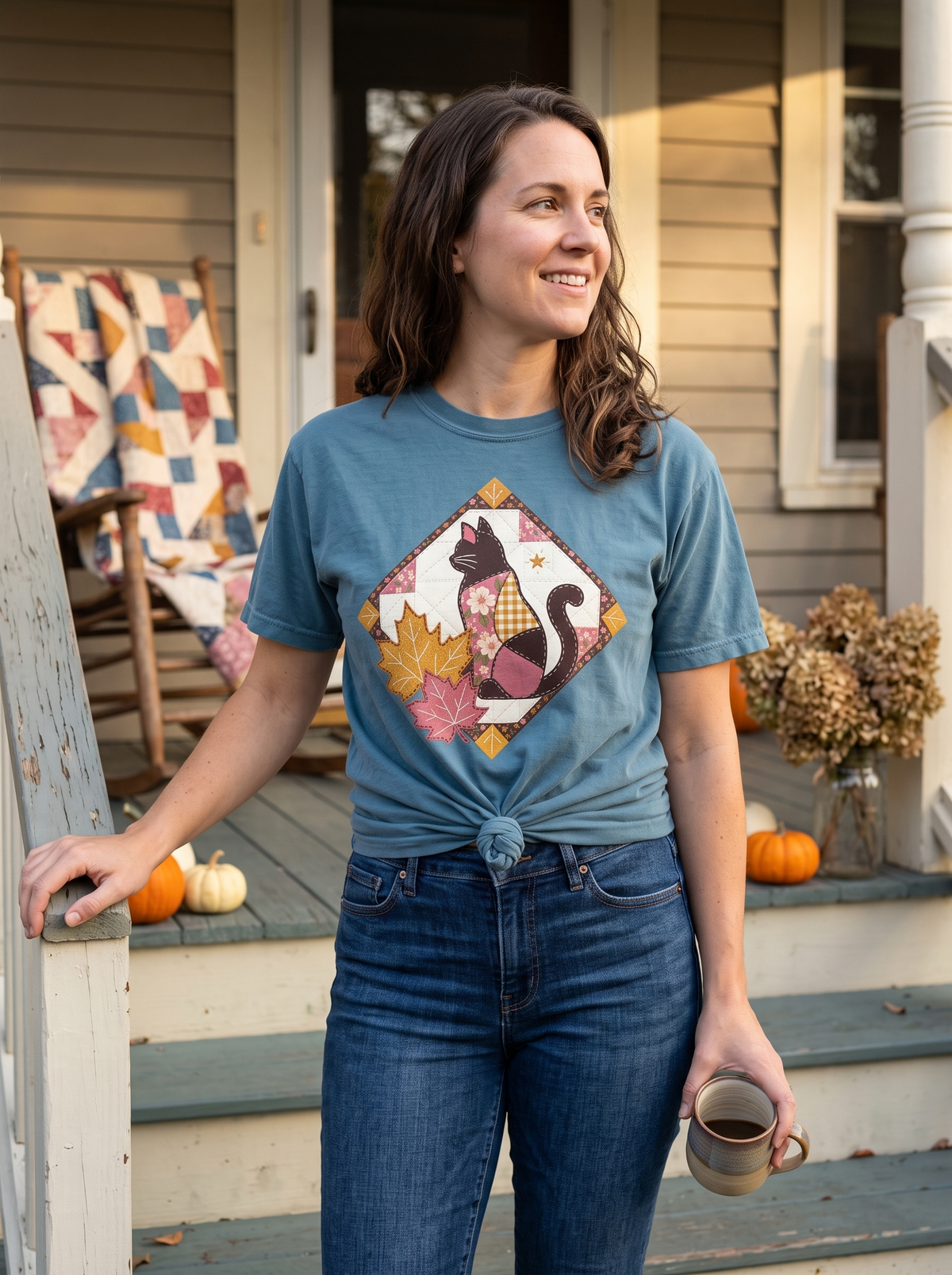 Woman in a Bay Comfort Colors tee with the patchwork quilt black cat print, knotted front hem, on autumn front porch steps with a mug and a quilt over the rail