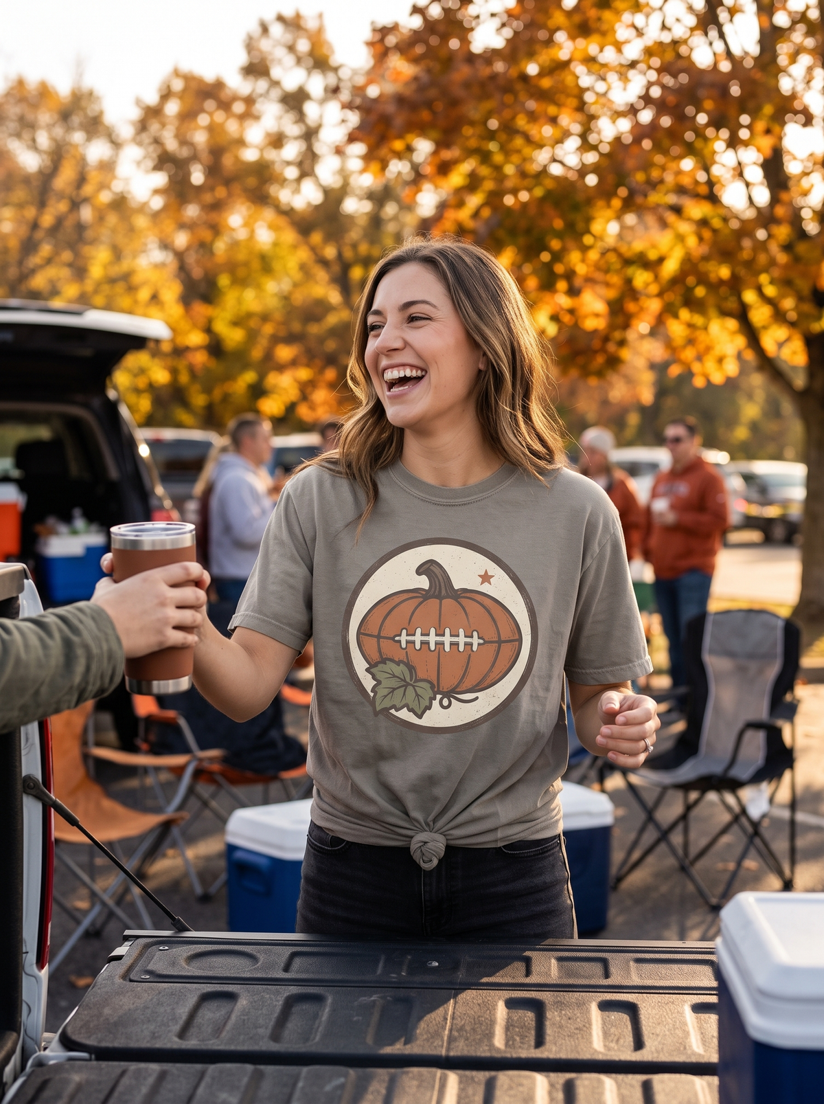 Woman in a Granite Comfort Colors tee with the vintage pumpkin football badge print, mid-laugh handing a drink across an open truck tailgate at an autumn stadium car park