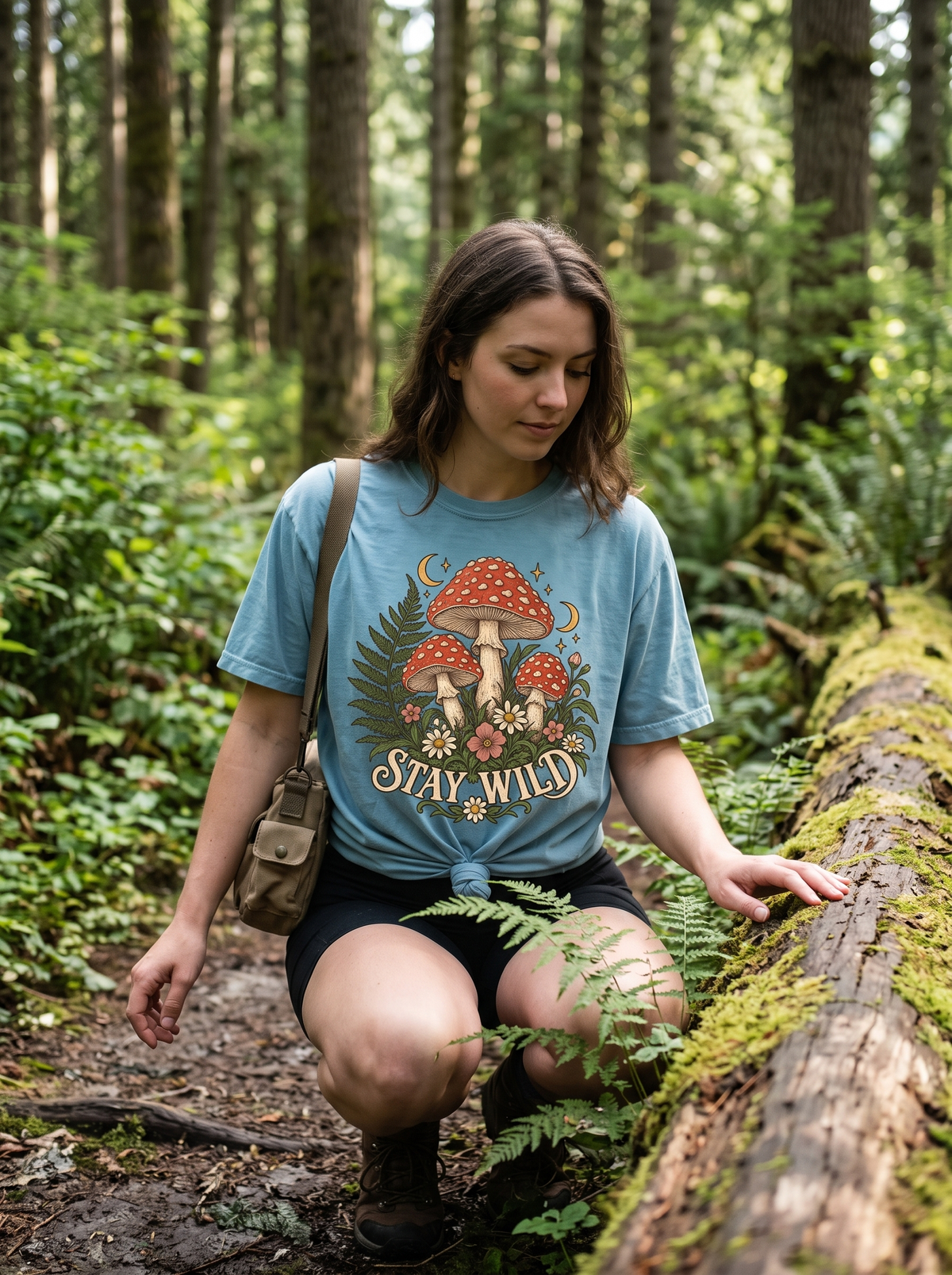 Woman in a Bay Comfort Colors tee with the Stay Wild fly agaric mushroom print, knotted front hem, crouched beside a mossy log on a damp woodland trail
