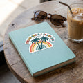 Palm Trees and 80 Degrees die-cut sticker applied to a linen journal beside sunglasses and an iced coffee
