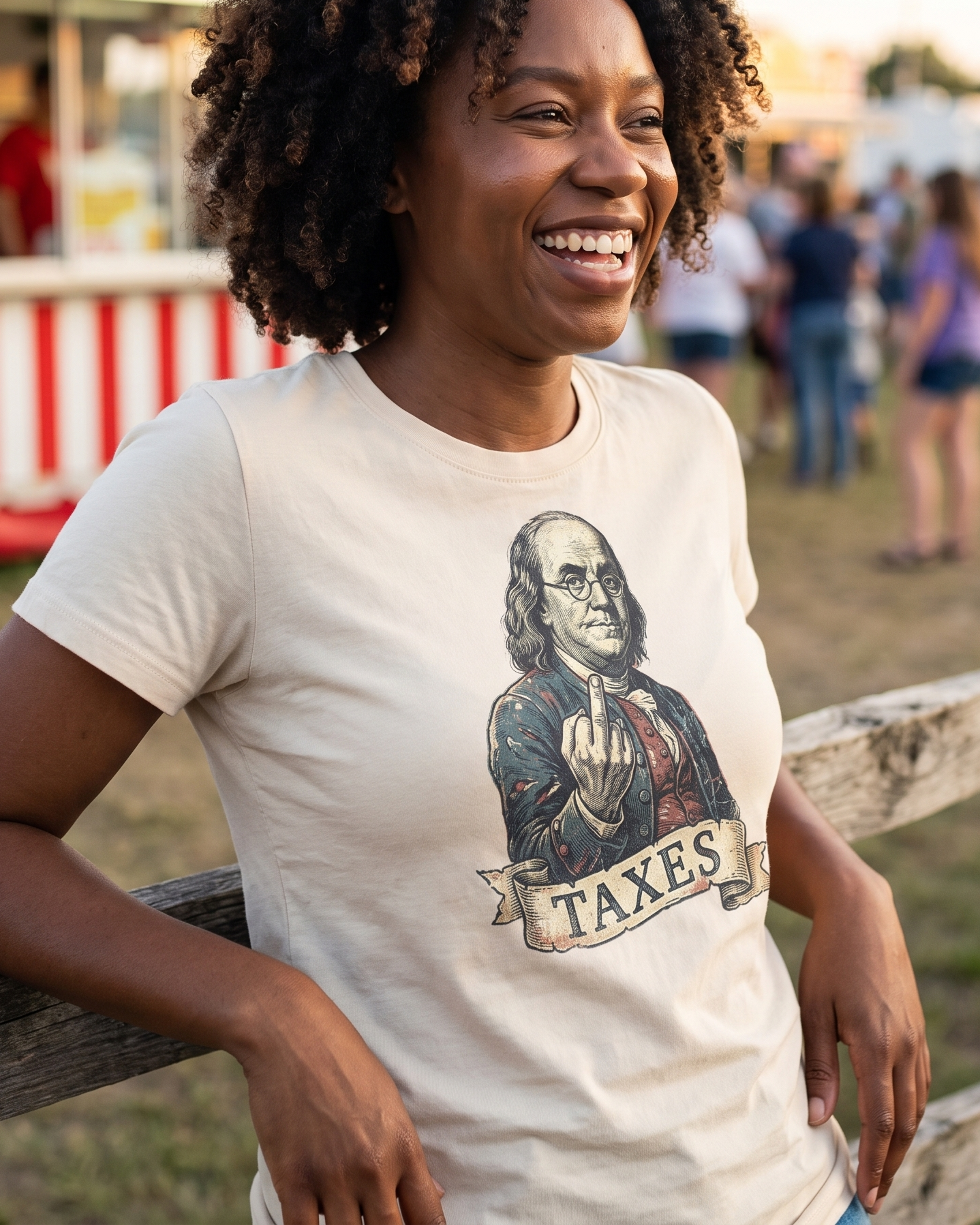 Woman laughing at a county fair fence in the Ben Franklin TAXES tee in Natural, engraved Franklin print and TAXES banner fully legible at mid chest, golden afternoon light