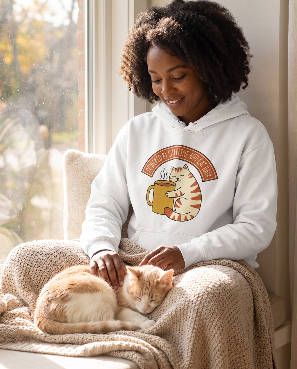 Woman on a window seat with a cat asleep in her lap in the Cat Coffee Powered by Caffeine and Cat Hair hoodie in White, print fully legible