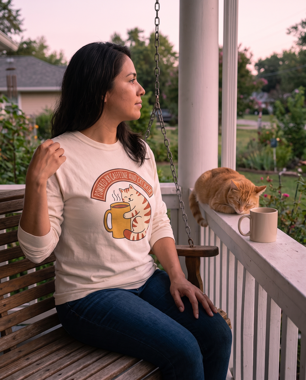 Woman on a porch swing at dusk in the Cat Coffee Powered by Caffeine and Cat Hair long sleeve in Ivory, print fully legible, cat asleep on the rail