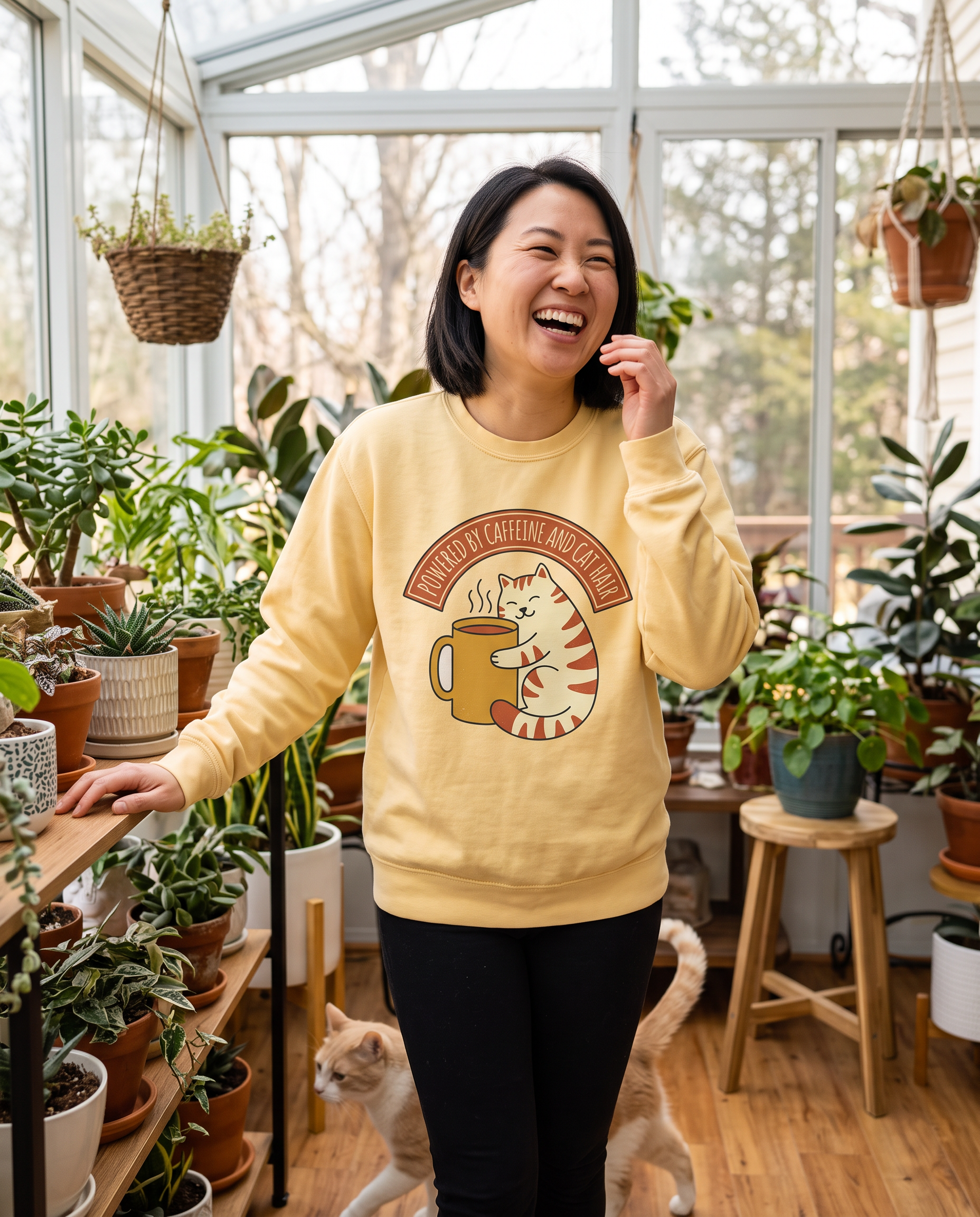 Woman laughing in a plant filled sunroom in the Cat Coffee Powered by Caffeine and Cat Hair sweatshirt in Butter, print fully legible