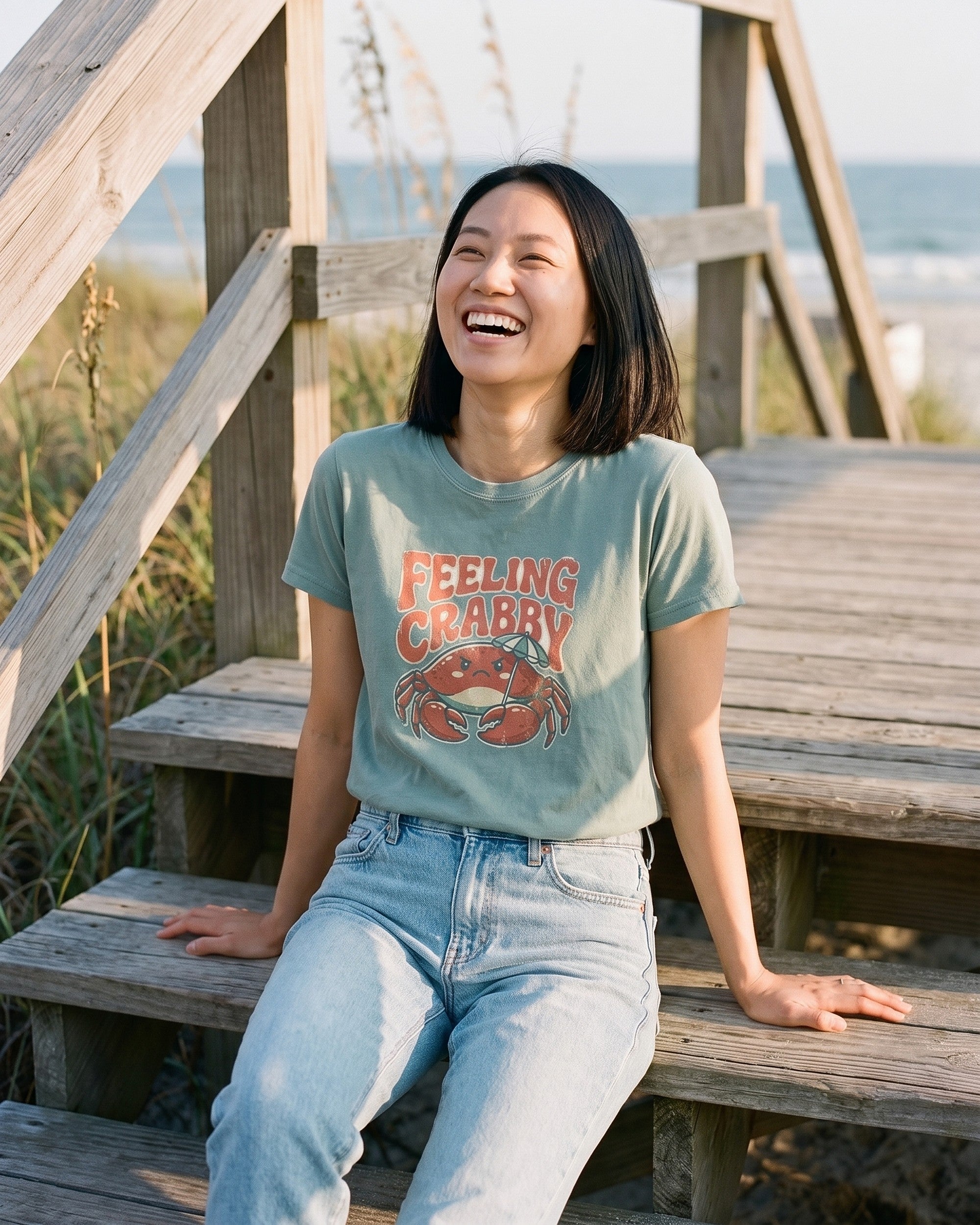 Woman laughing on weathered dune steps wearing a sage green Feeling Crabby tee with a grumpy red crab holding a striped beach umbrella. AI generated model imagery, no real person photographed