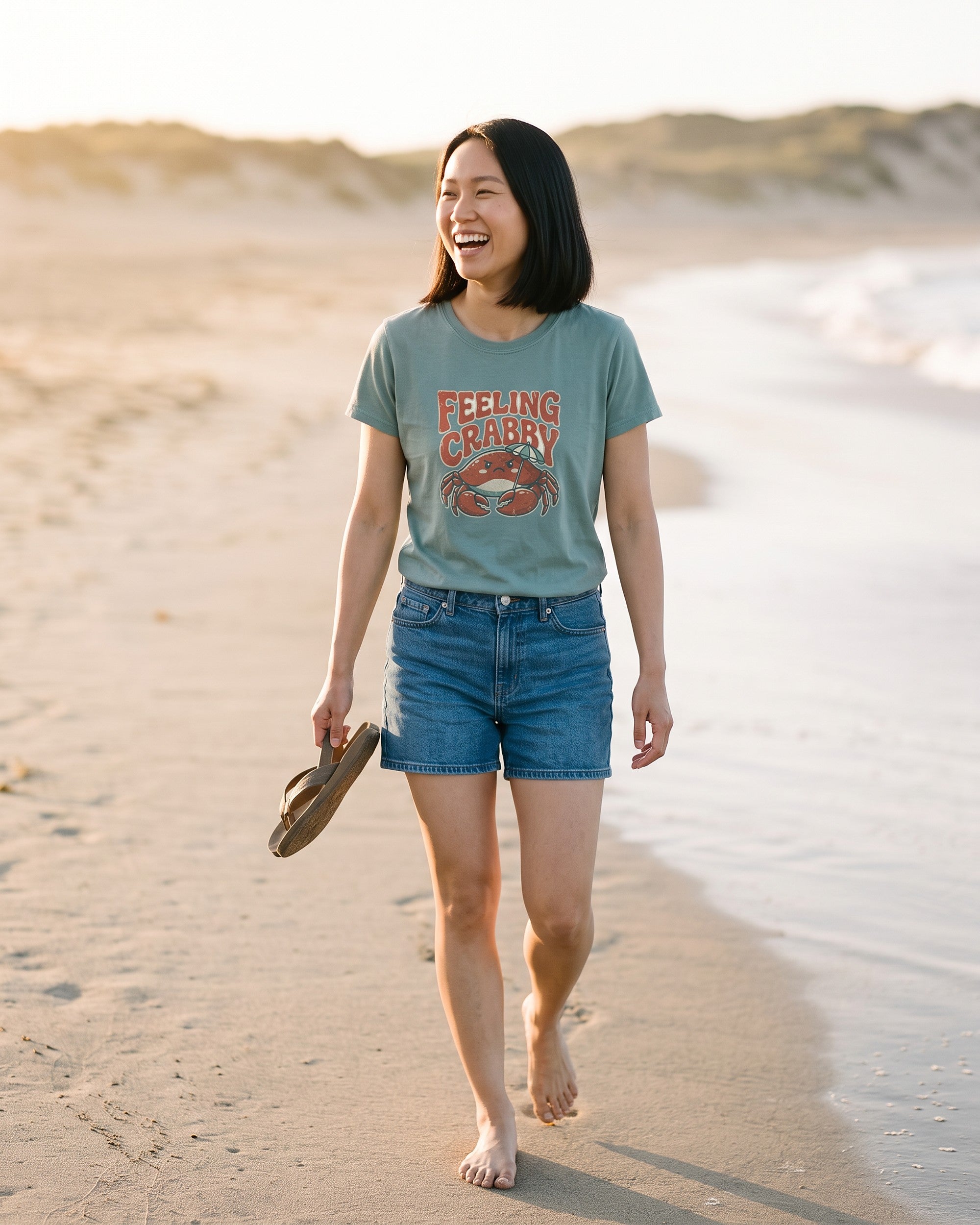 Woman walking barefoot along the tide line carrying her sandals, wearing a sage green Feeling Crabby tee with a grumpy red crab. AI generated model imagery, no real person photographed