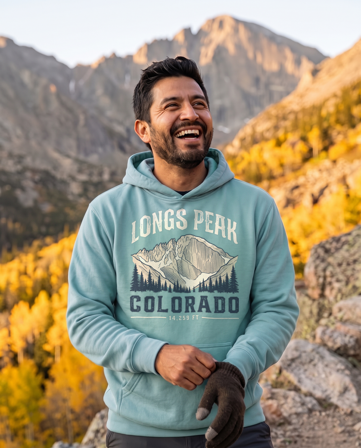 Laughing man pulling on a glove below a granite peak and golden aspens, wearing a Seafoam Longs Peak Colorado hoodie