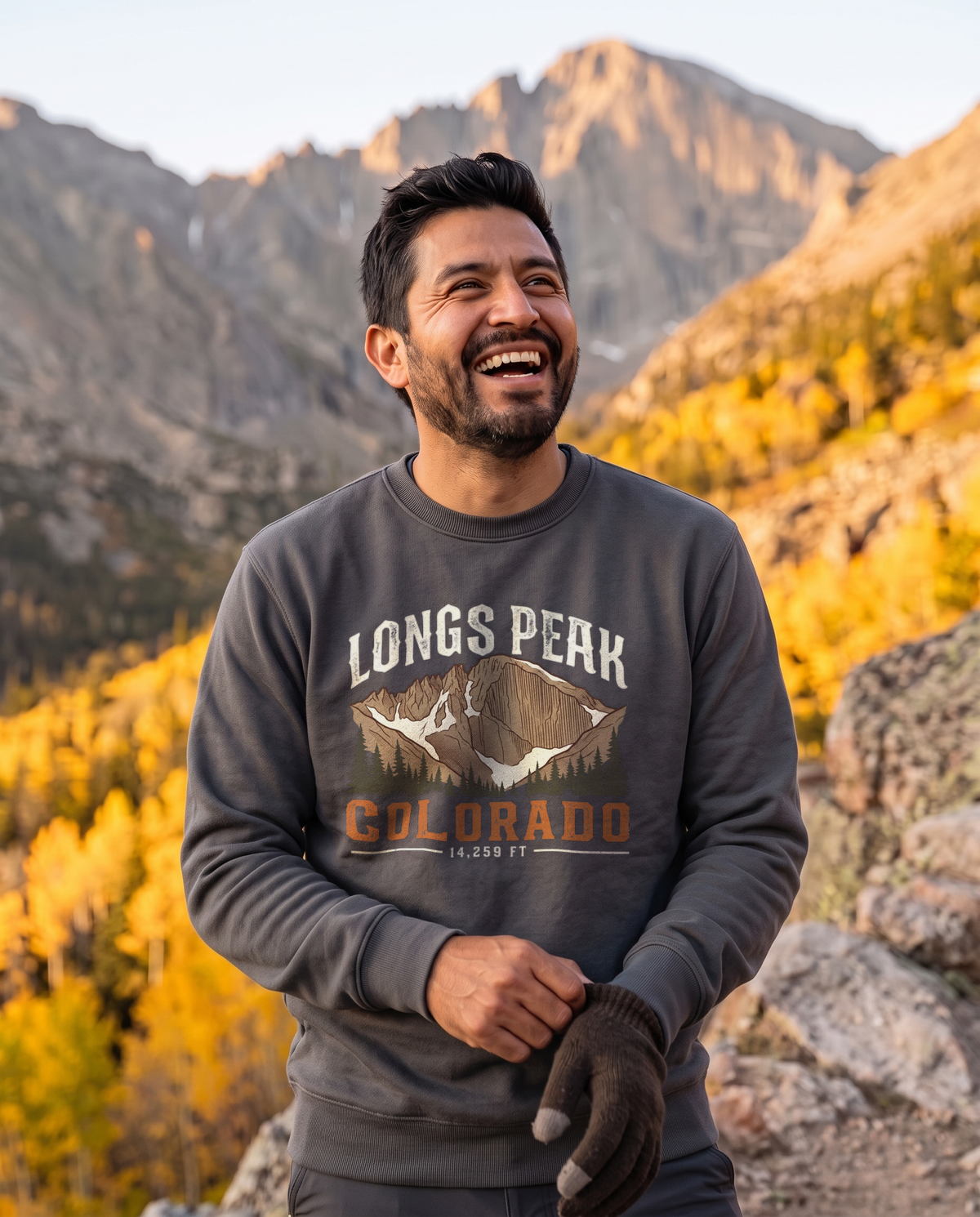 Laughing man pulling on a glove below a granite peak and golden aspens, wearing a Smoke grey Longs Peak Colorado crewneck