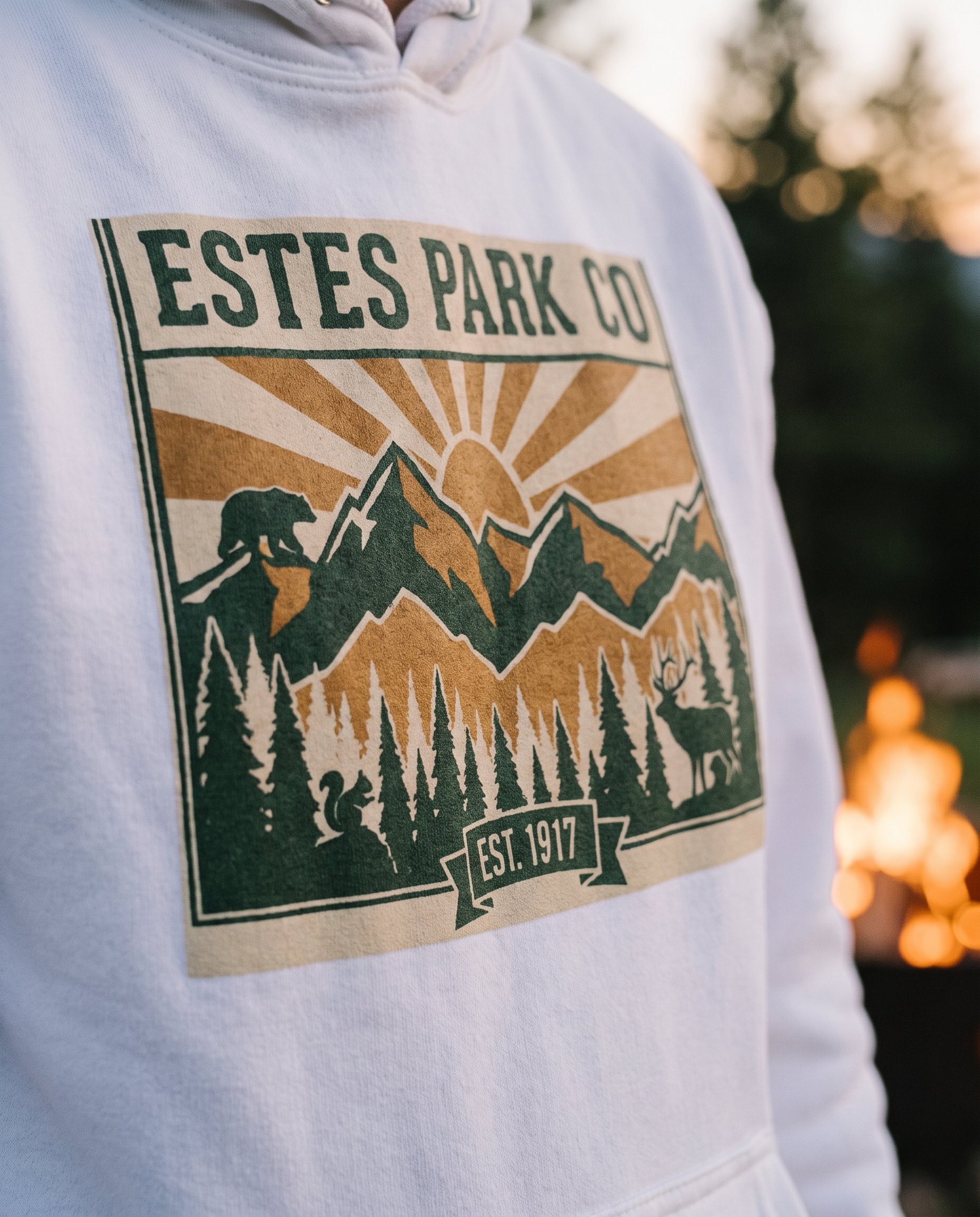 Estes Park CO hidden wildlife badge close-up on a white hoodie by the campfire, bear elk and squirrel design, EST. 1917