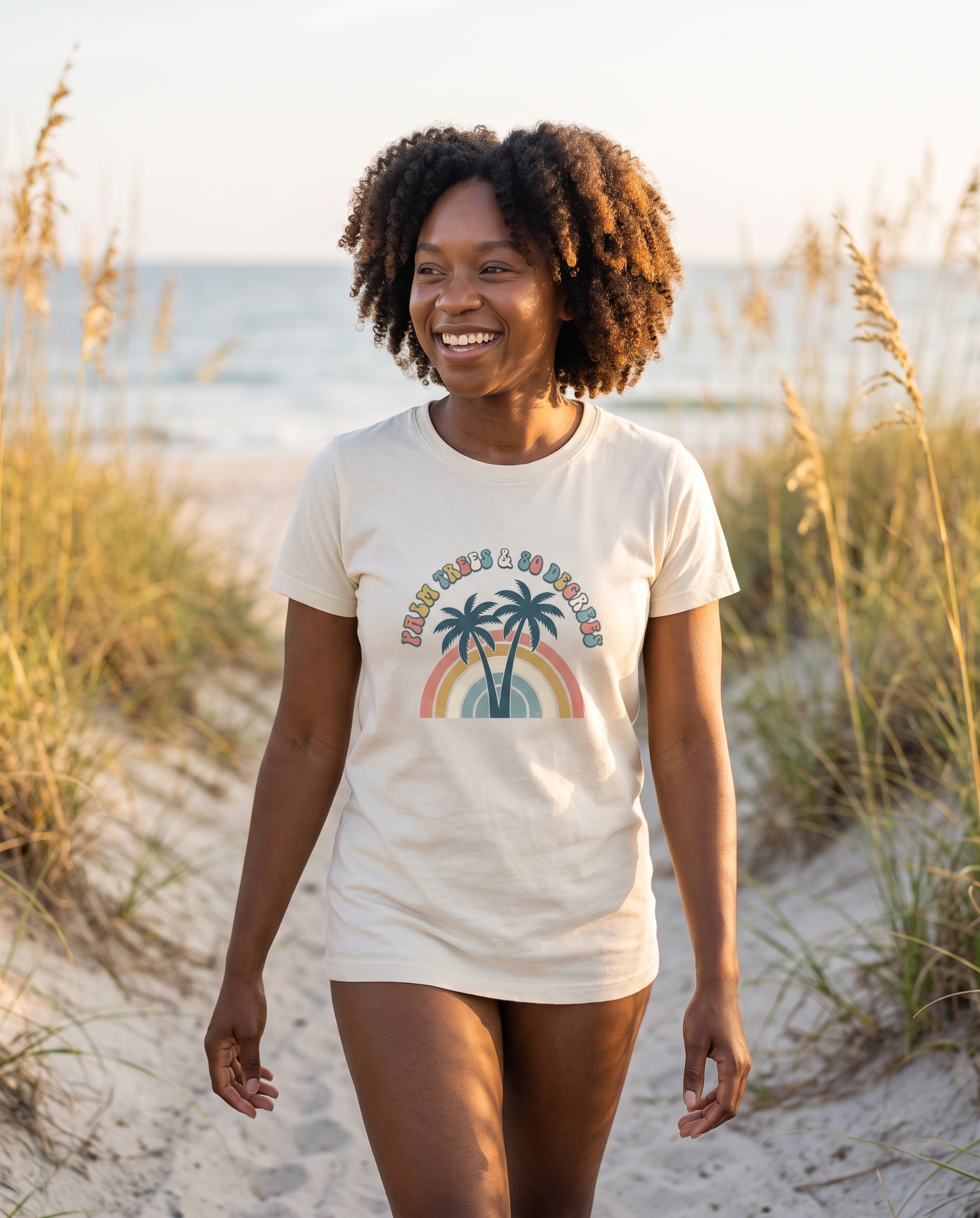Woman walking a sea oat dune path in the Palm Trees and 80 Degrees tee in Ivory, warm afternoon light, retro rainbow palm design visible
