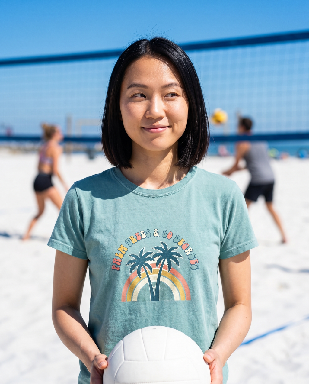 Woman holding a volleyball at a beach volleyball net wearing the Palm Trees and 80 Degrees tee in Bay, retro palm rainbow print on the chest