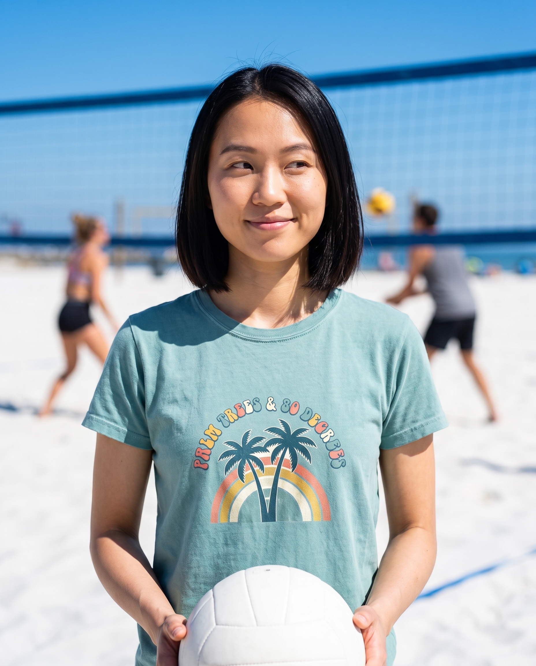Woman holding a volleyball at a beach volleyball net wearing the Palm Trees and 80 Degrees tee in Bay, retro palm rainbow print on the chest