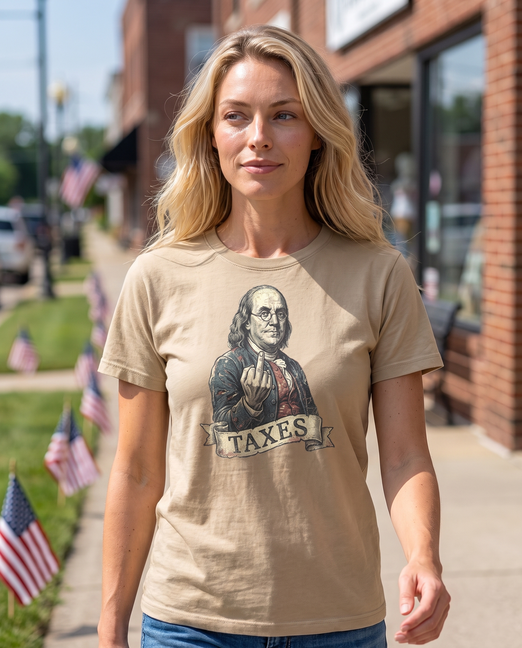 Woman strolling a small town July 4th parade sidewalk in the Ben Franklin TAXES tee in Sand, engraved Franklin print visible, flags on the lawns