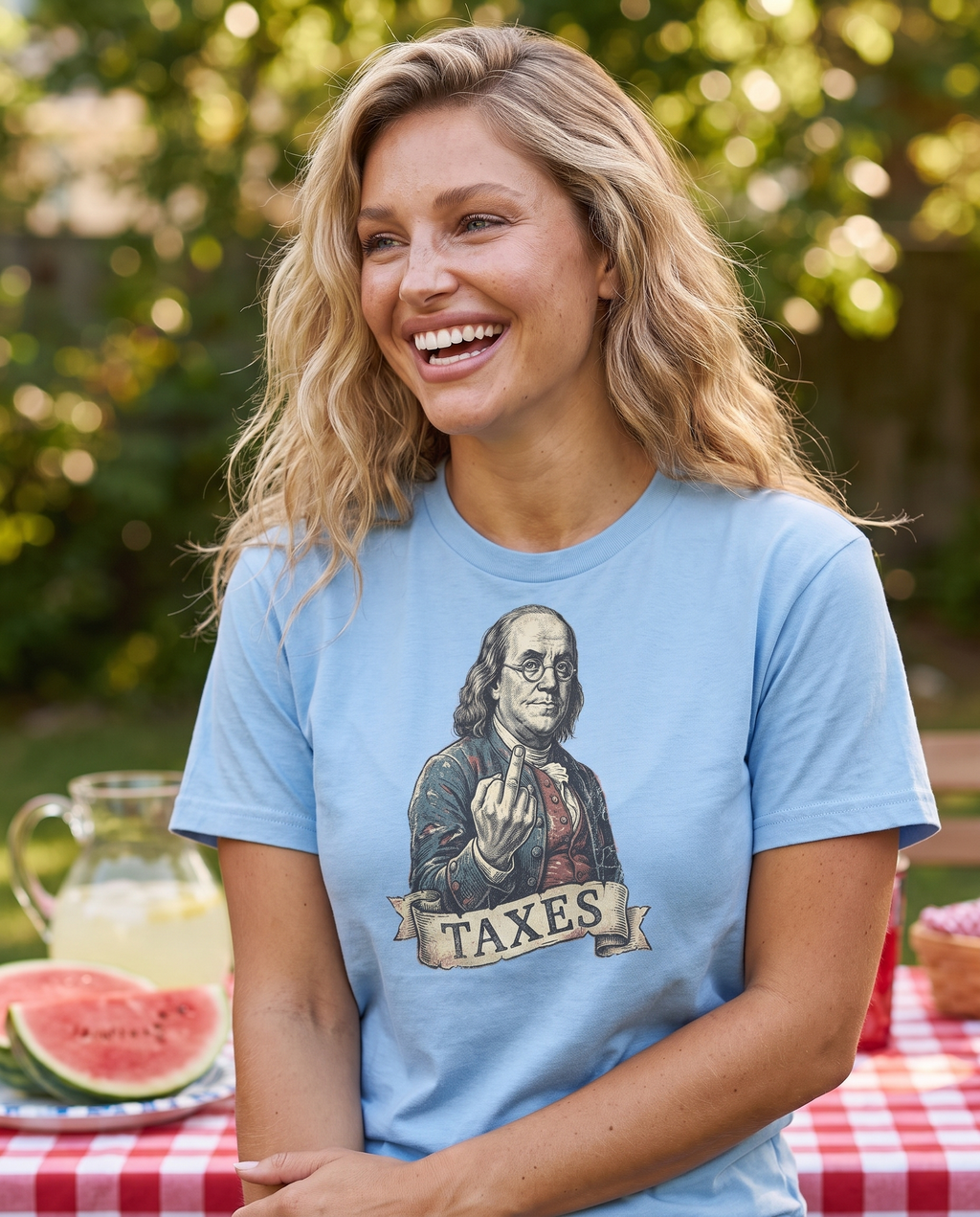 Woman laughing at a backyard July 4th picnic in the Ben Franklin TAXES tee in Carolina Blue, engraved Franklin print visible, watermelon table behind