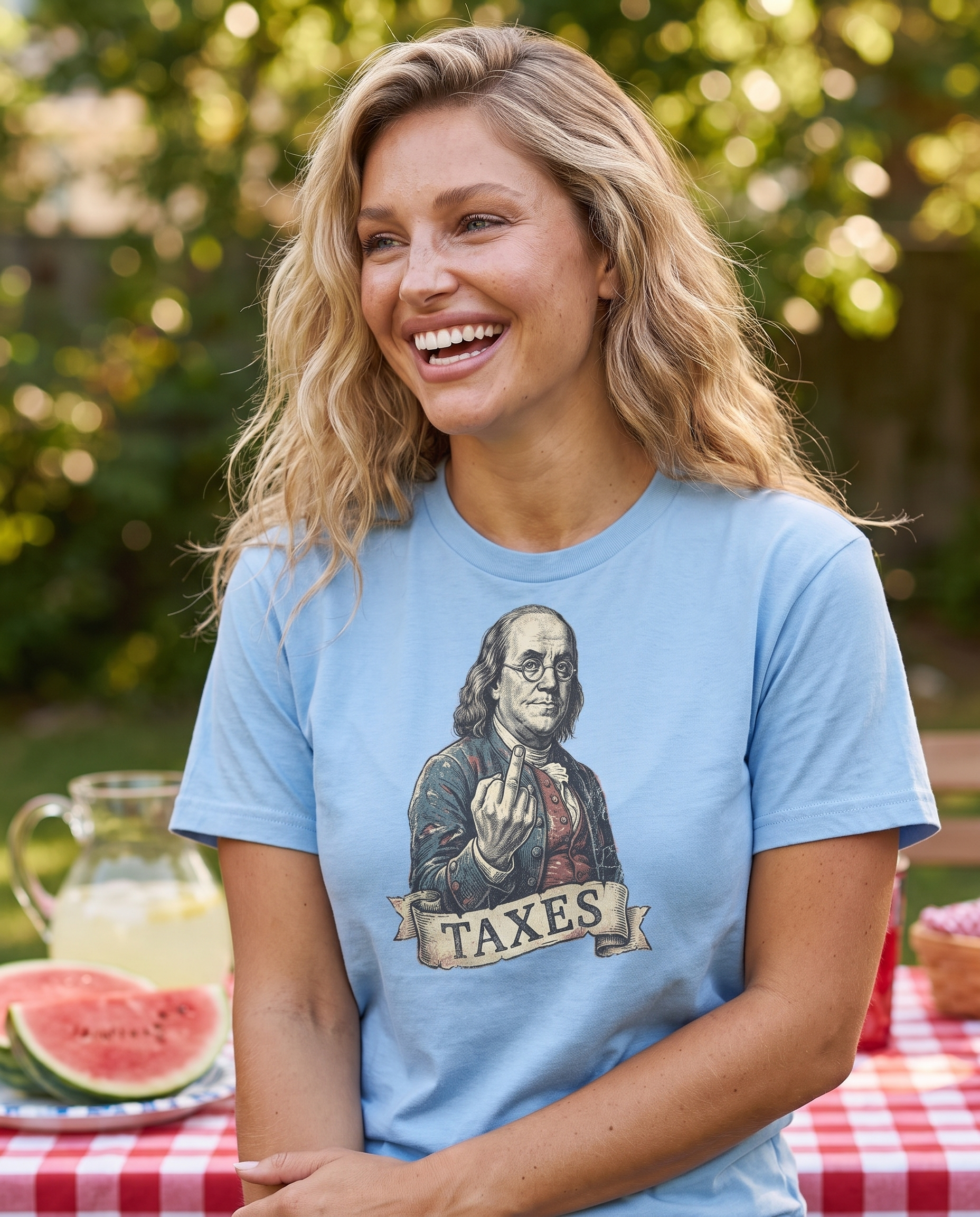 Woman laughing at a backyard July 4th picnic in the Ben Franklin TAXES tee in Carolina Blue, engraved Franklin print visible, watermelon table behind