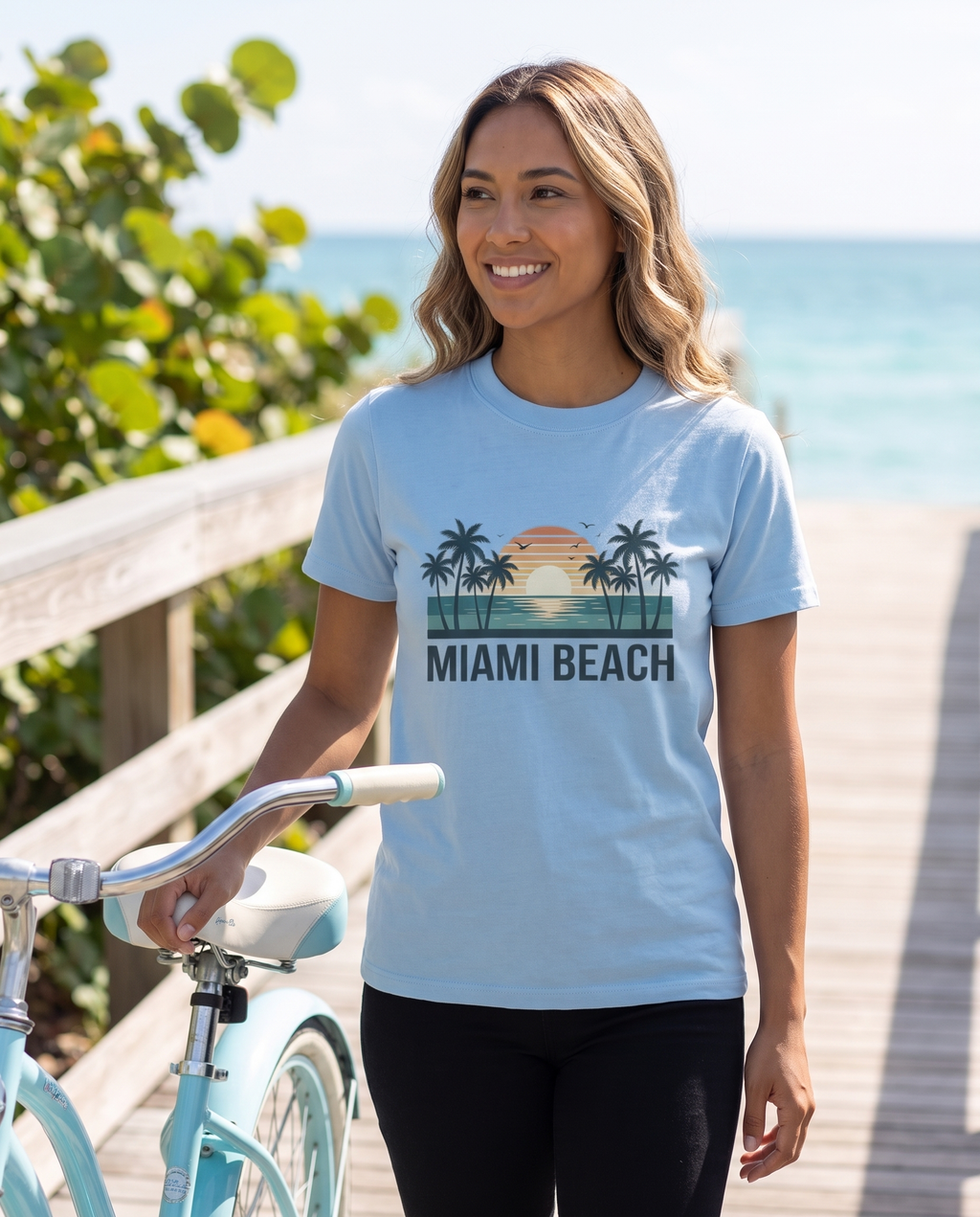 Woman walking a pastel beach cruiser on a Miami boardwalk in the Miami Beach retro tee in Carolina Blue, retro sunset design visible