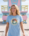 Woman walking toward a pastel lifeguard tower in the South Beach Miami badge tee in Carolina Blue, badge visible, bright beach light