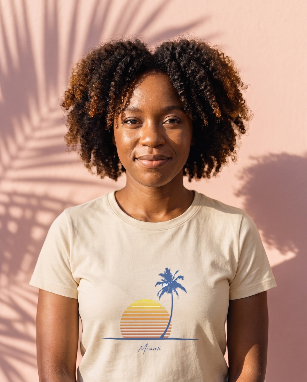 Woman at a pastel pink art deco wall in the Retro Miami Palm Sunset tee in Natural, single palm striped sunset print with Miami script at mid chest
