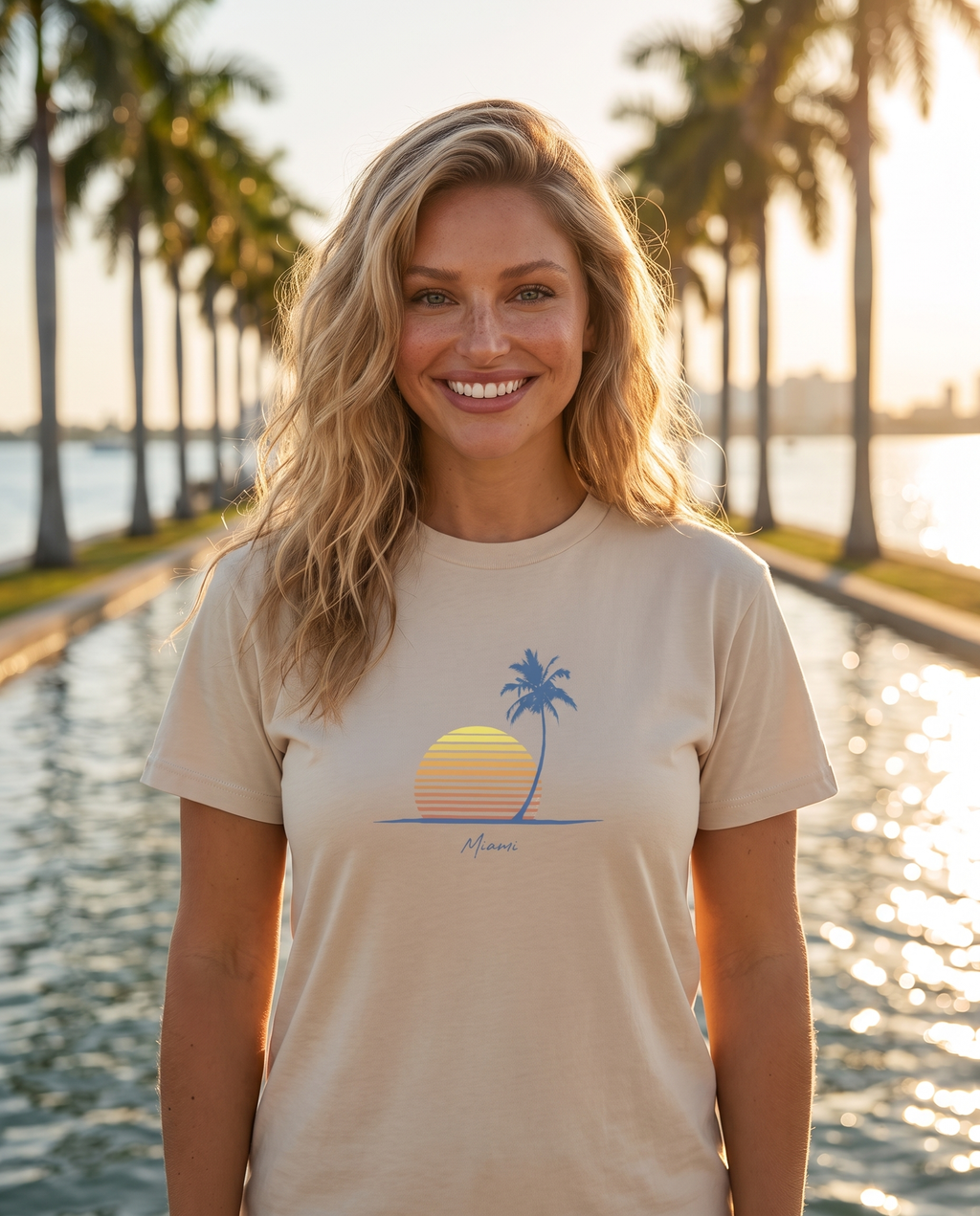 Woman on a palm lined causeway at golden hour in the Retro Miami Palm Sunset tee in Sand, single palm striped sunset print with Miami script at mid chest