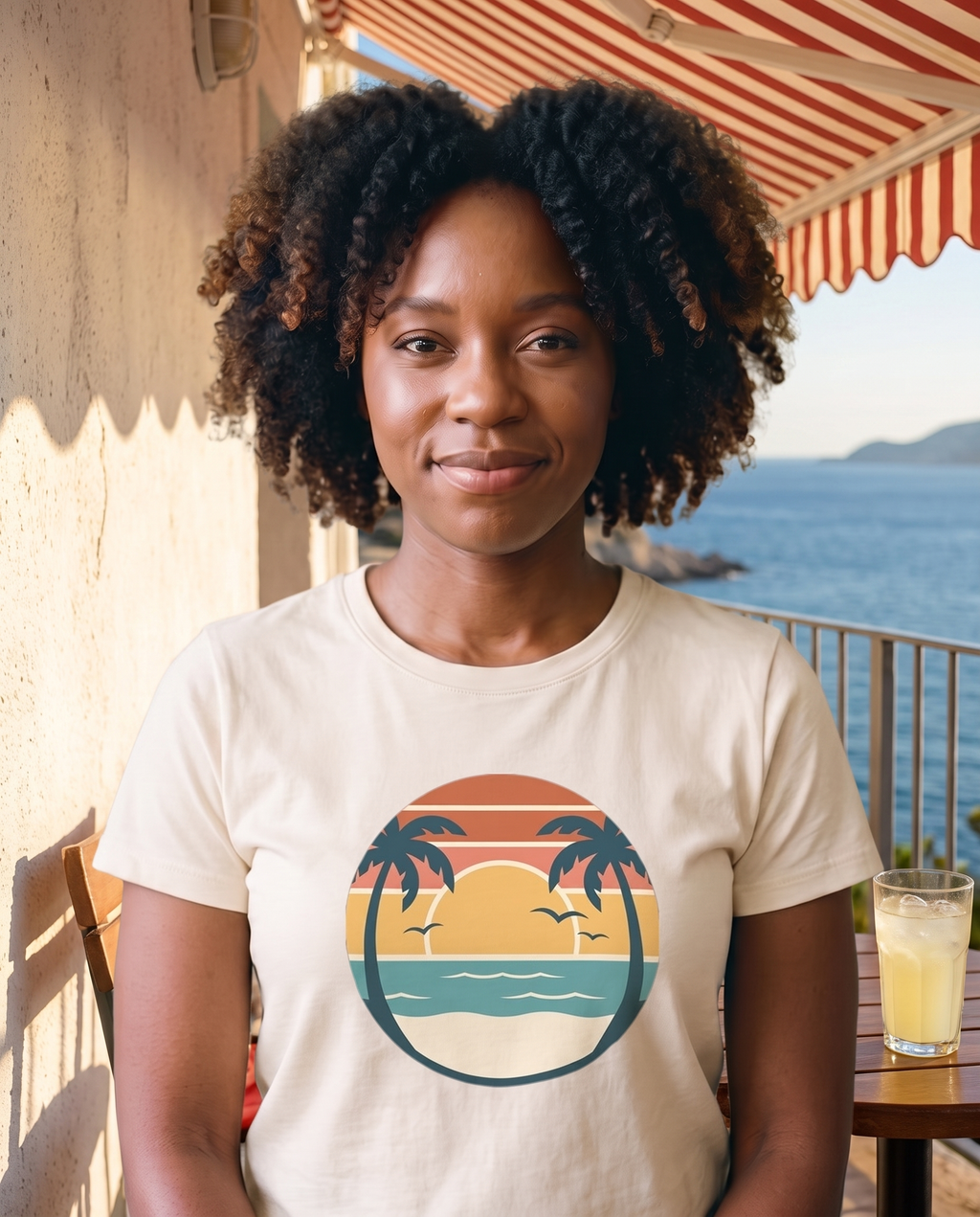 Woman at a seaside cafe terrace in the Retro Sunset Circle tee in Natural, striped sunset circle badge with palms at mid chest