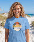 Woman on a white sand dune path with sea oats in the Retro Sunset Circle tee in Carolina Blue, striped sunset circle badge with palms at mid chest