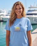Woman at a Miami marina in the Retro Miami Palm Sunset tee in Carolina Blue, single palm striped sunset print with Miami script at mid chest