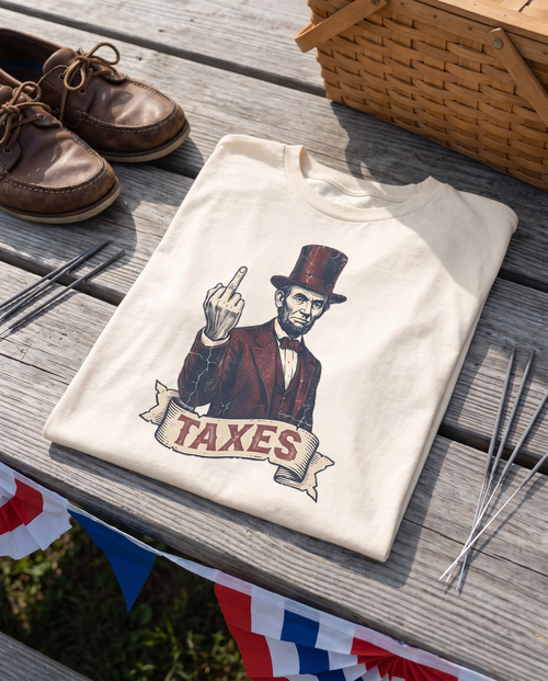 Cream garment-dyed cotton tee folded face up on the weathered grey planks of an outdoor picnic table, its chest print showing a vintage engraved Abraham Lincoln in a stovepipe hat above a ribbon banner reading TAXES. Brown leather boat shoes, a woven picnic basket, sparklers and red white and blue paper bunting sit softly out of focus around it in warm afternoon light.