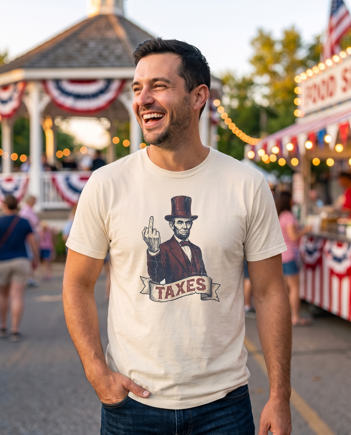 Man in his early thirties laughing at an outdoor summer street fair, wearing a cream garment-dyed cotton tee printed with a vintage engraved Abraham Lincoln in a stovepipe hat above a ribbon banner reading TAXES. Blurred food stalls, string lights and a white gazebo sit behind him in golden hour light.