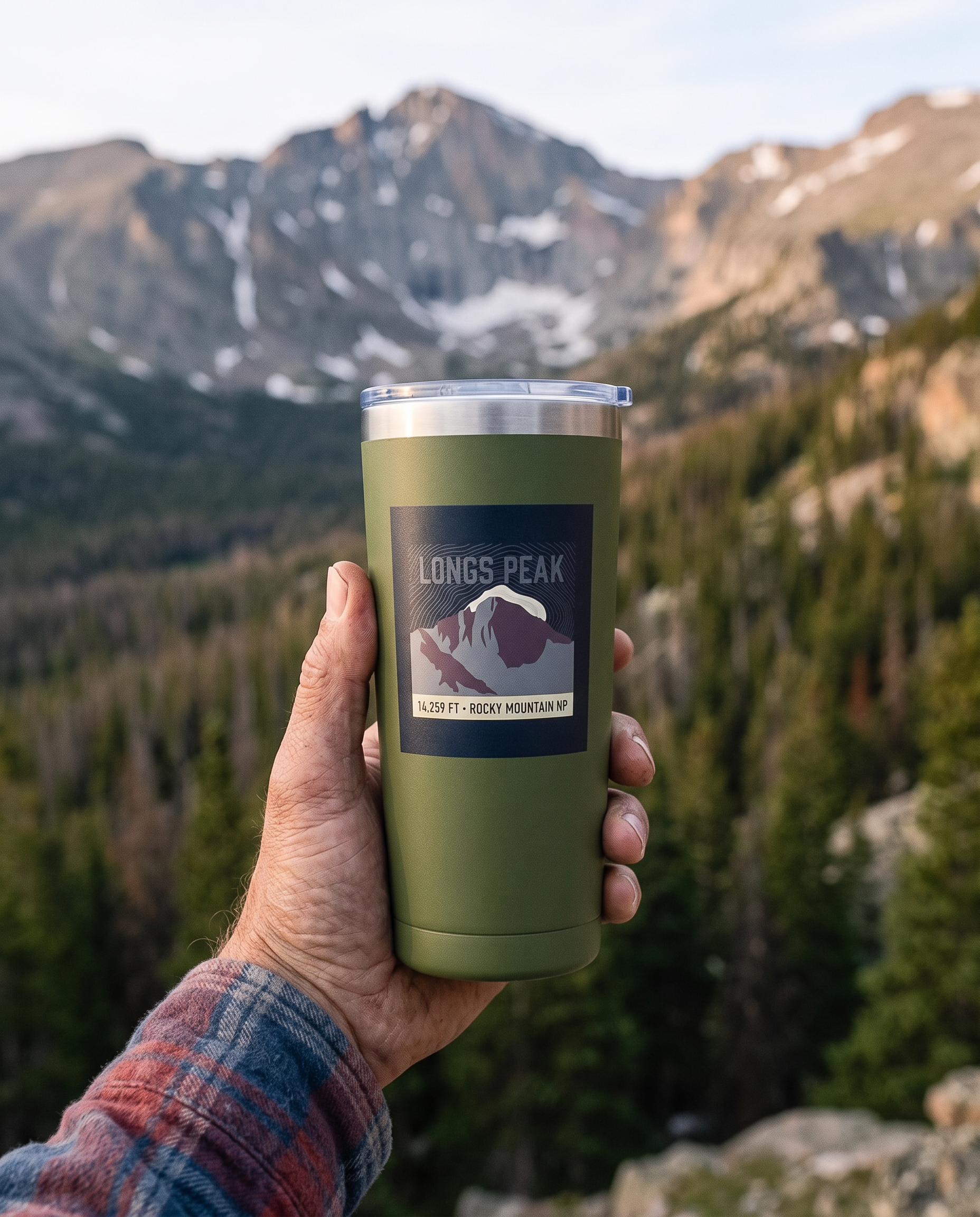 A weathered hand in a rolled flannel sleeve holding a matte olive green 20oz insulated tumbler at a high alpine overlook at sunrise, the tumbler printed with a dark navy topographic badge reading LONGS PEAK above a mauve mountain with a white snow cap and the line 14,259 FT ROCKY MOUNTAIN NP. A conifer valley and distant granite ridge lie below.