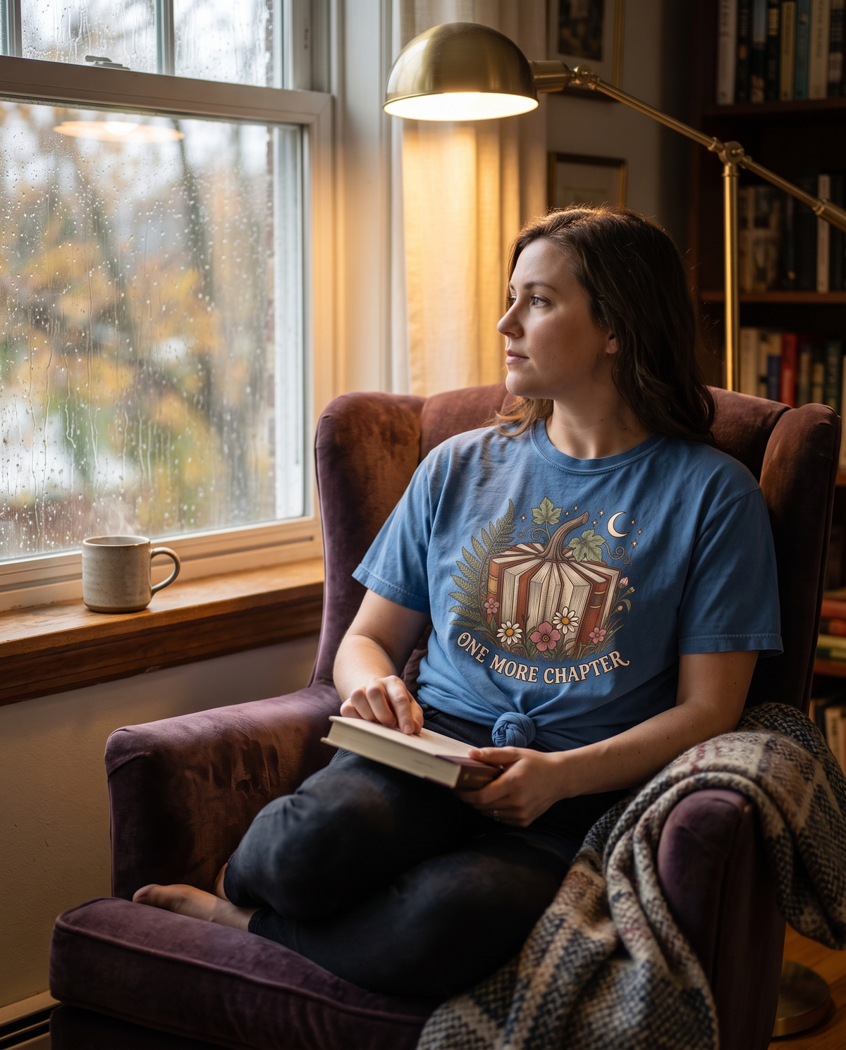 Woman in a blue Book Lover One More Chapter tee reading in an armchair by a rainy window