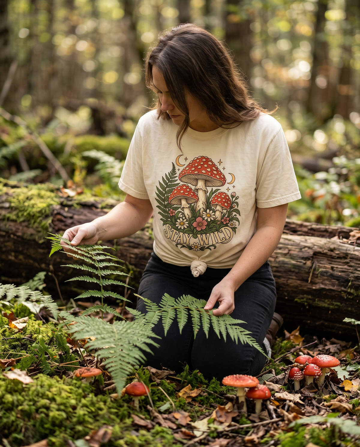 Woman in a cream Stay Wild Mushroom tee kneeling on a mossy woodland bank among ferns