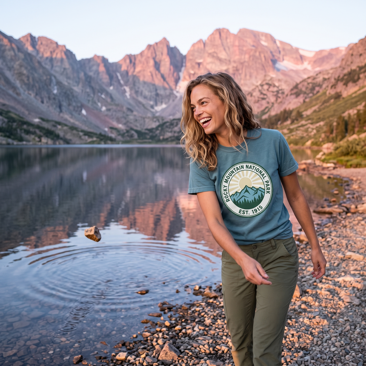 A woman walks a rocky lakeshore below pink alpine peaks in a blue tee printed with a round ROCKY MOUNTAIN NATIONAL PARK EST. 1915 badge.