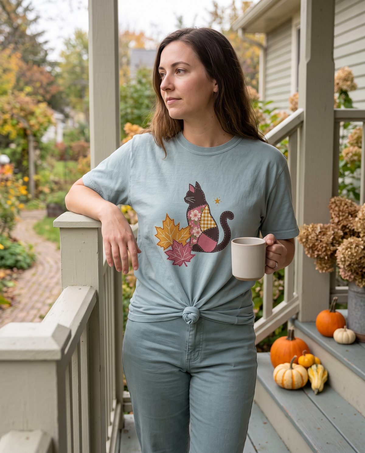 Woman in a pale blue quilted Black Cat tee leaning on an autumn porch rail with a mug