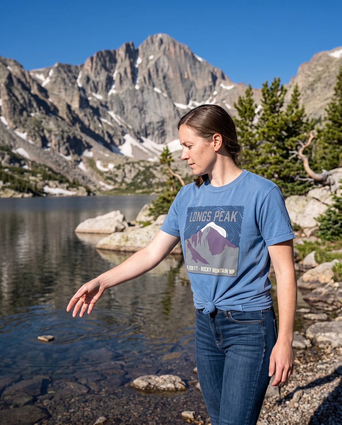 Woman in a dusty blue Longs Peak tee at the shore of an alpine lake below the granite summit