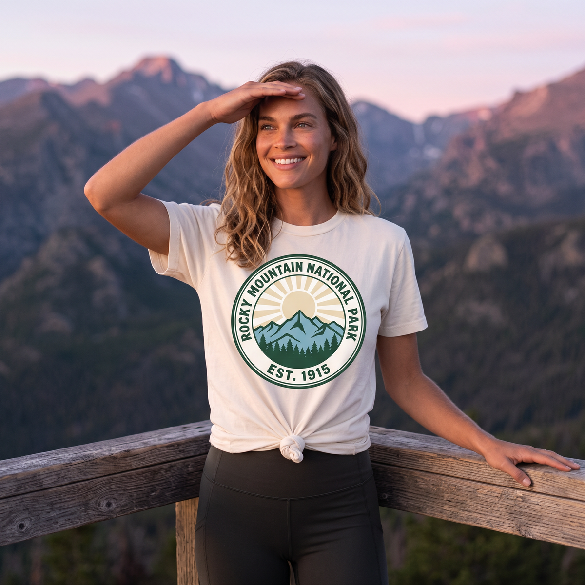 A woman shades her eyes at a wooden overlook rail with mountains behind, in a cream tee knotted at the hem with a ROCKY MOUNTAIN NATIONAL PARK EST. 1915 badge.