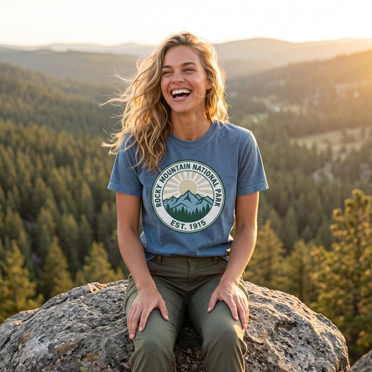 A woman laughs sitting on a granite boulder above a pine valley at sunset, in a blue tee with a round ROCKY MOUNTAIN NATIONAL PARK EST. 1915 badge.