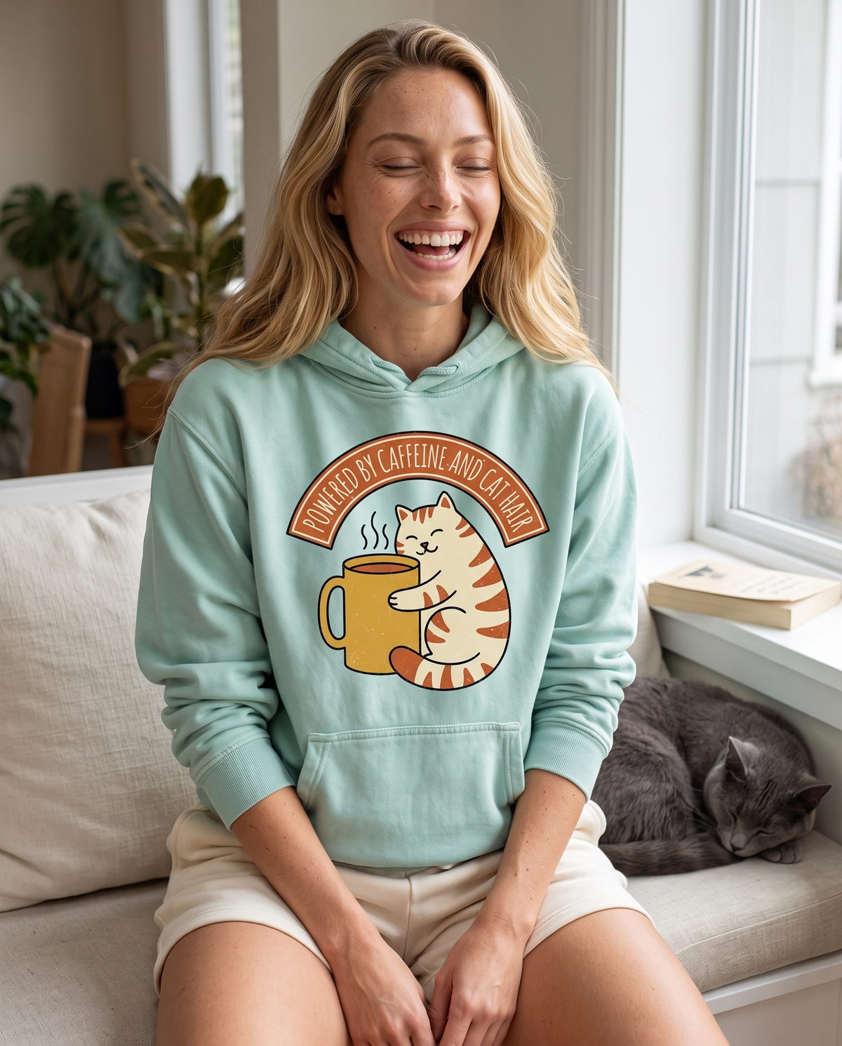 A woman laughs on a window seat beside a sleeping grey cat, in a mint hoodie printed POWERED BY CAFFEINE AND CAT HAIR above a cat hugging a yellow mug.