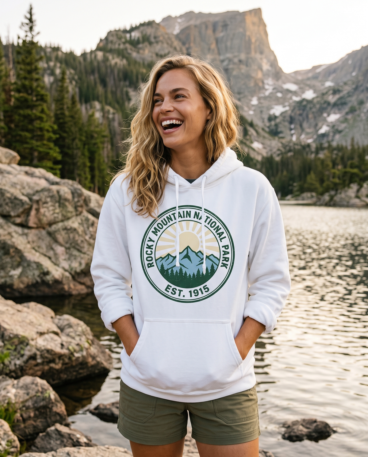 A woman stands on rocks beside an alpine lake with hands in her pockets, in a white hoodie printed with a ROCKY MOUNTAIN NATIONAL PARK EST. 1915 badge.