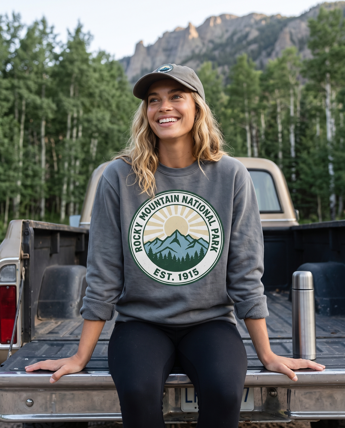 A woman in a cap sits on a pickup tailgate beside a steel flask, in a grey crewneck sweatshirt with a ROCKY MOUNTAIN NATIONAL PARK EST. 1915 badge.