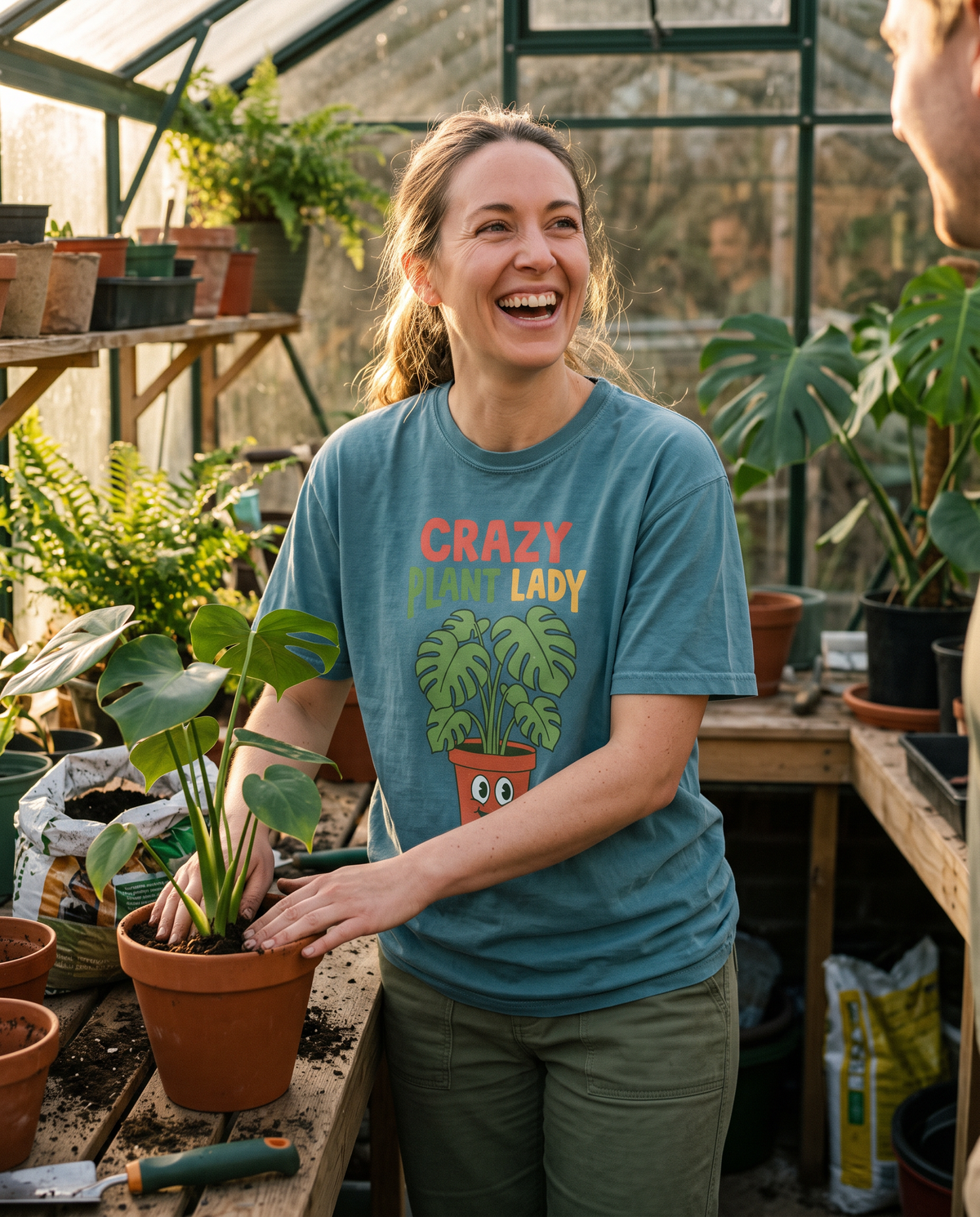 Woman laughing in a greenhouse while potting a monstera, wearing a Crazy Plant Lady tee, lifestyle photograph