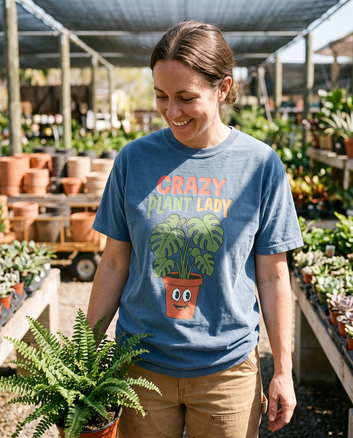 Woman in a Blue Jean Crazy Plant Lady tee carrying a fern down a plant nursery aisle, lifestyle photograph