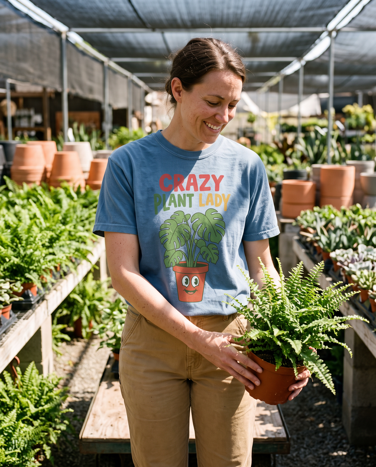 Woman in a Blue Jean Crazy Plant Lady tee holding a potted fern under nursery shade cloth, lifestyle photograph
