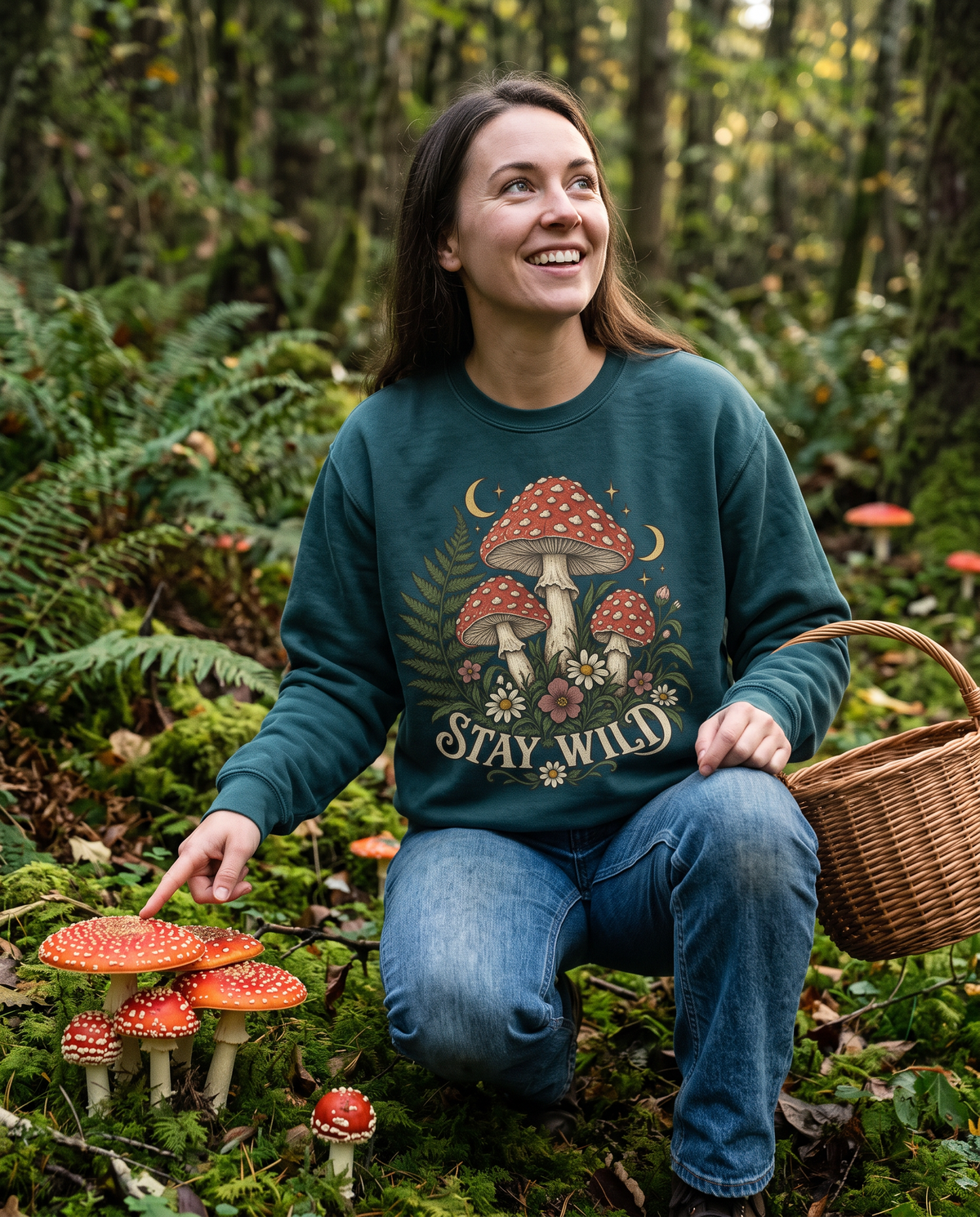 Woman in a Blue Spruce Mushroom crewneck sweatshirt crouched on a mossy forest floor beside red-capped toadstools, grinning, a wicker foraging basket at her hip.