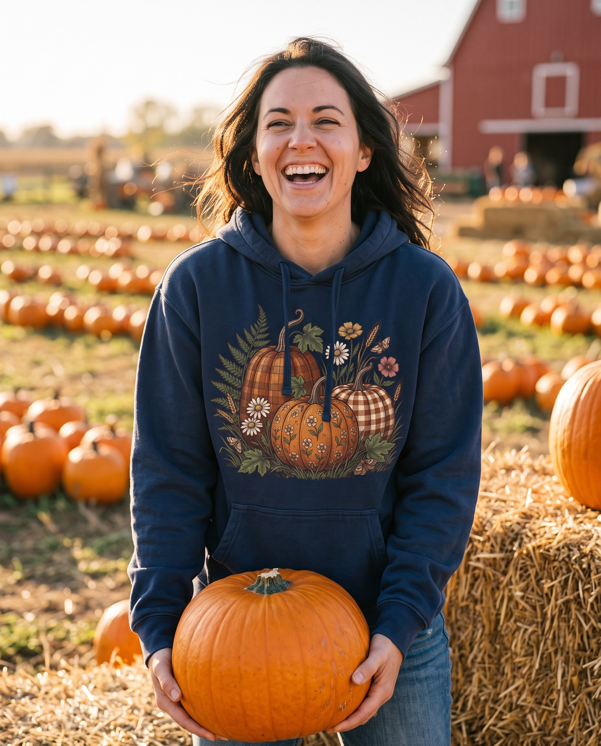 Woman in a True Navy Pumpkin hoodie lifting a fat orange pumpkin off a hay bale at a pumpkin patch, mid-laugh, in low warm harvest sun.