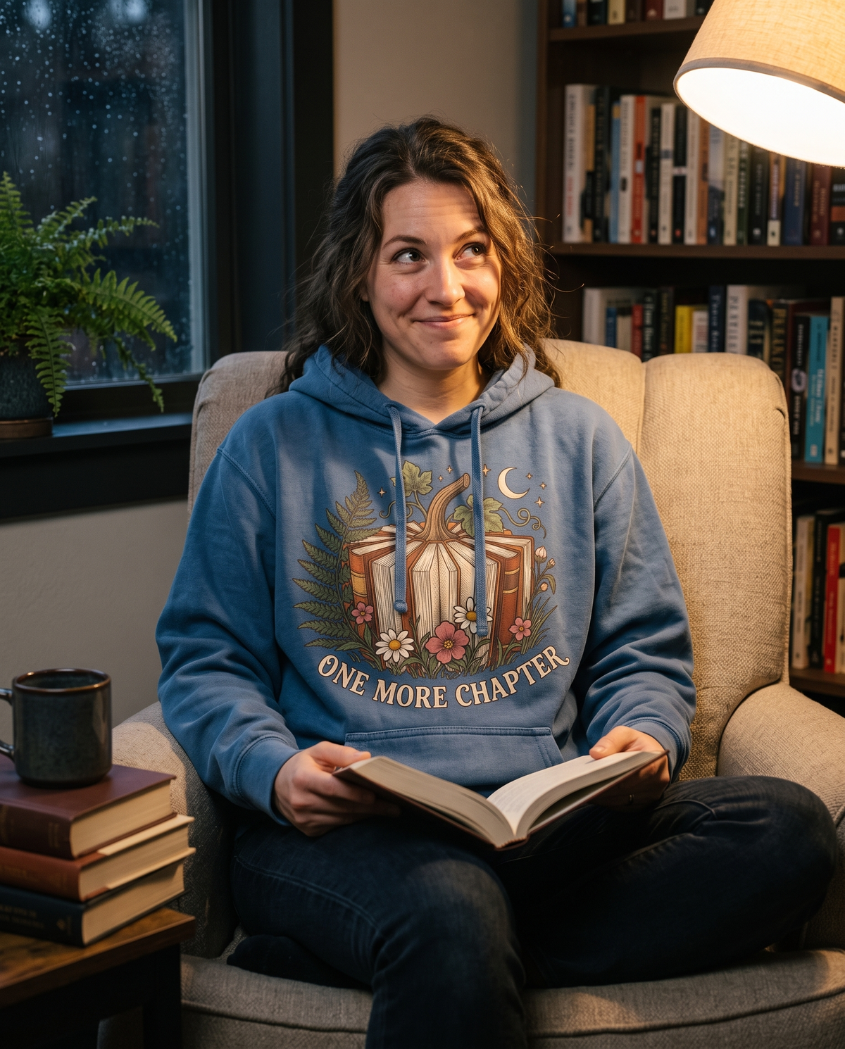 Woman in a Denim Book Lover hoodie curled in an armchair holding an open hardback, glancing up with a grin, a mug on the side table under warm lamplight.