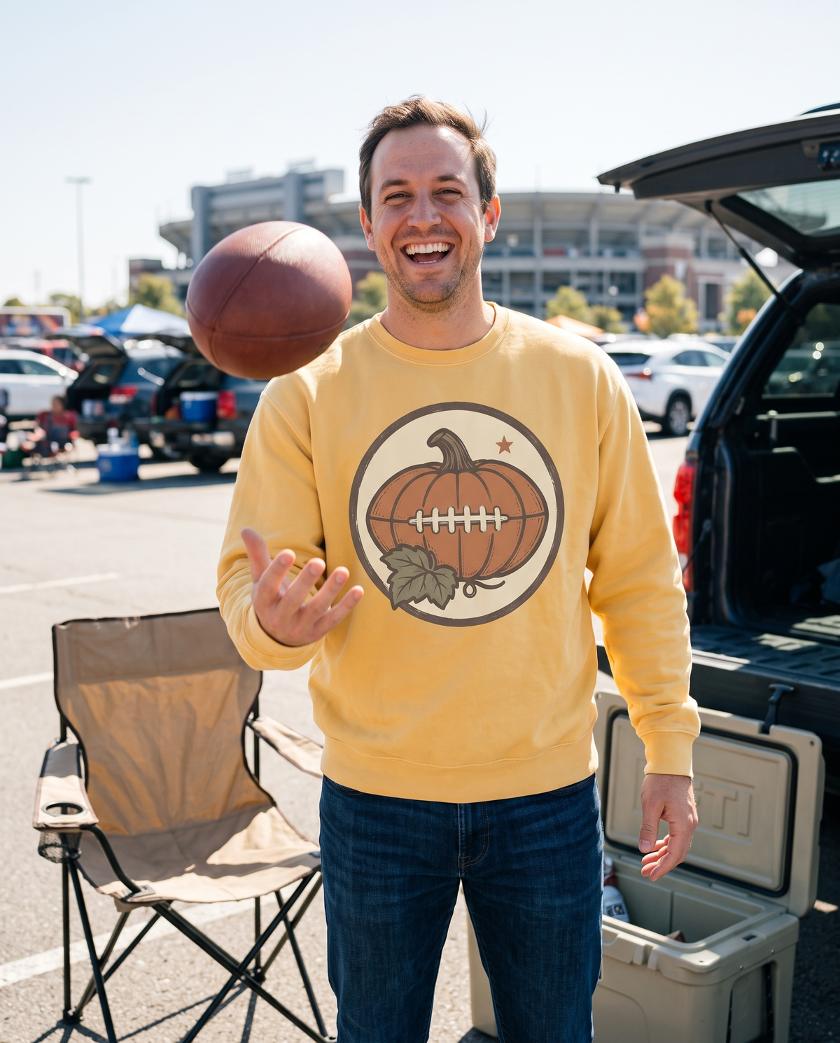 Man in a Butter Pumpkin Football crewneck sweatshirt at a tailgate tossing a football underhand toward the camera, laughing, a folding chair and cooler beside him.