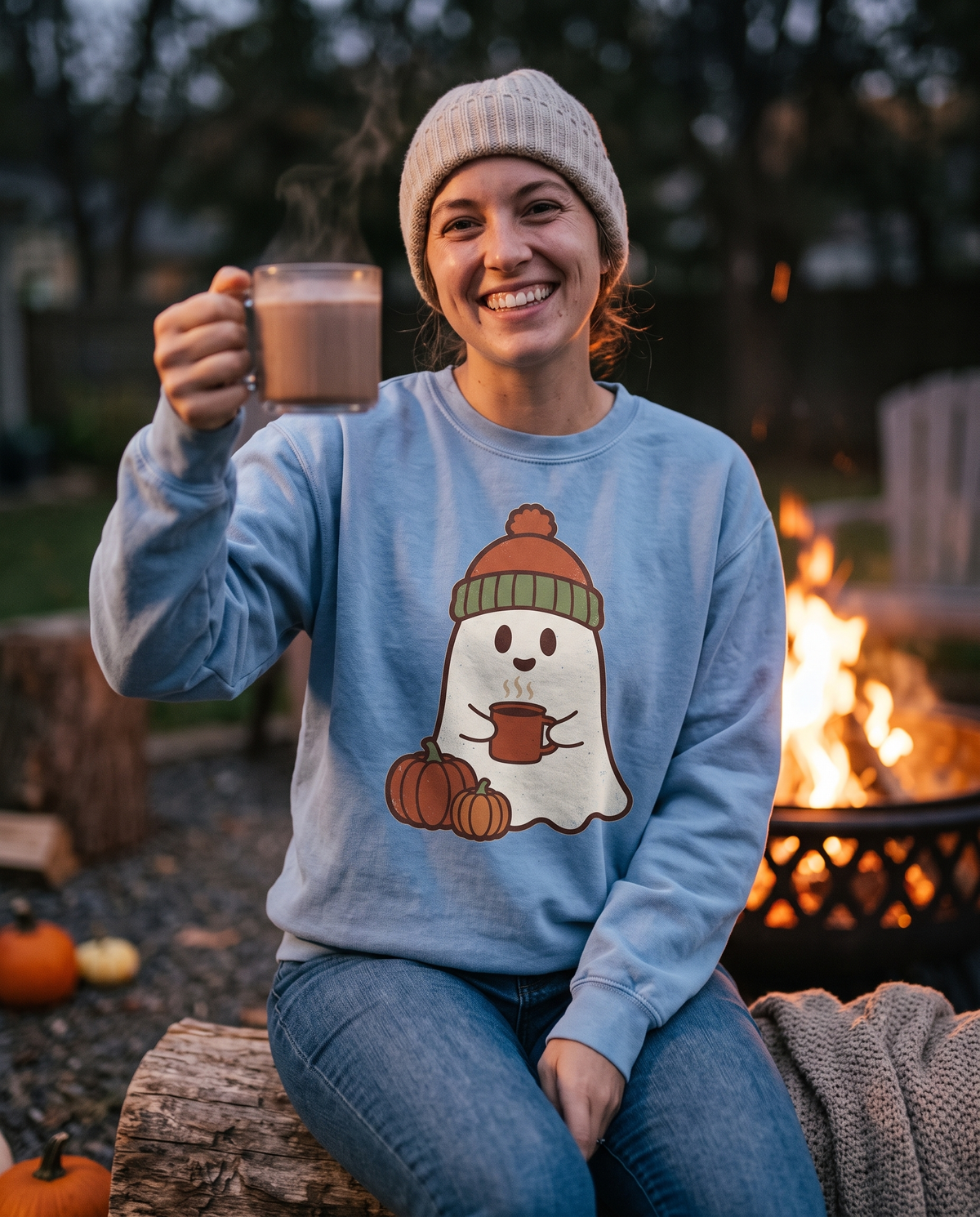 Woman in a Chambray Retro Ghost crewneck sweatshirt and a knitted beanie sitting on a log by a fire pit, raising a cocoa mug in a toast toward the camera, grinning.